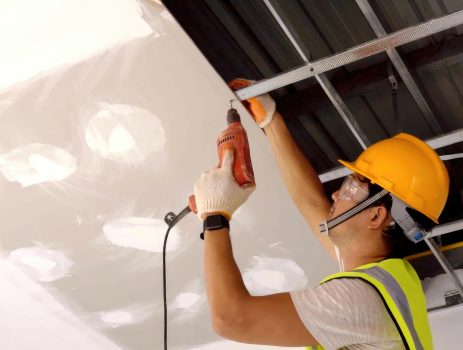 Asian construction worker in safety clothing and work gloves is fastening the drywall ceiling to the metal frame using an electric screwdriver on the ceiling covere